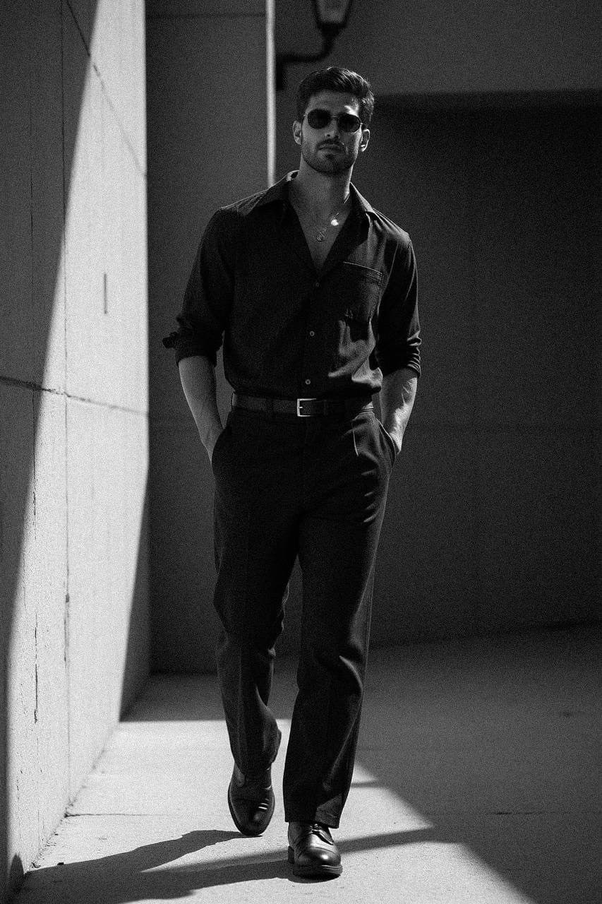 A man walks along a massive architectural wall, wearing an oversized black shirt and high-waisted trousers, casting deep shadows in strong light.