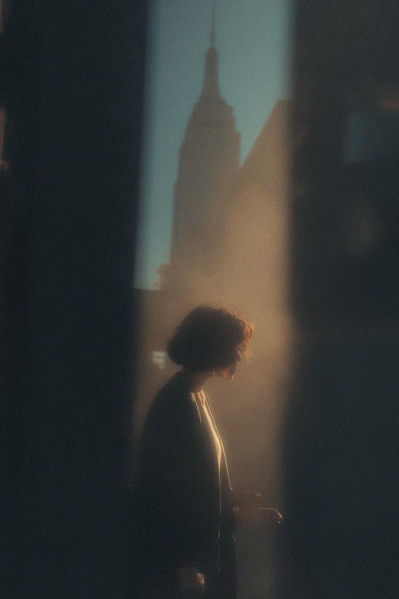 A woman in profile stands silhouetted by warm golden sunlight, creating a dramatic effect with fine film grain and atmospheric haze.