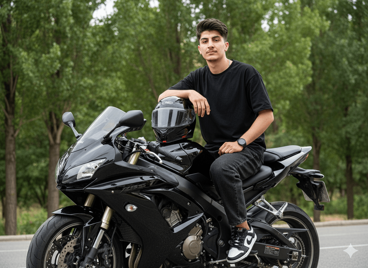 A young person sitting on a shiny black sports motorcycle in an outdoor setting surrounded by green trees, wearing a loose black T-shirt and dark jeans.